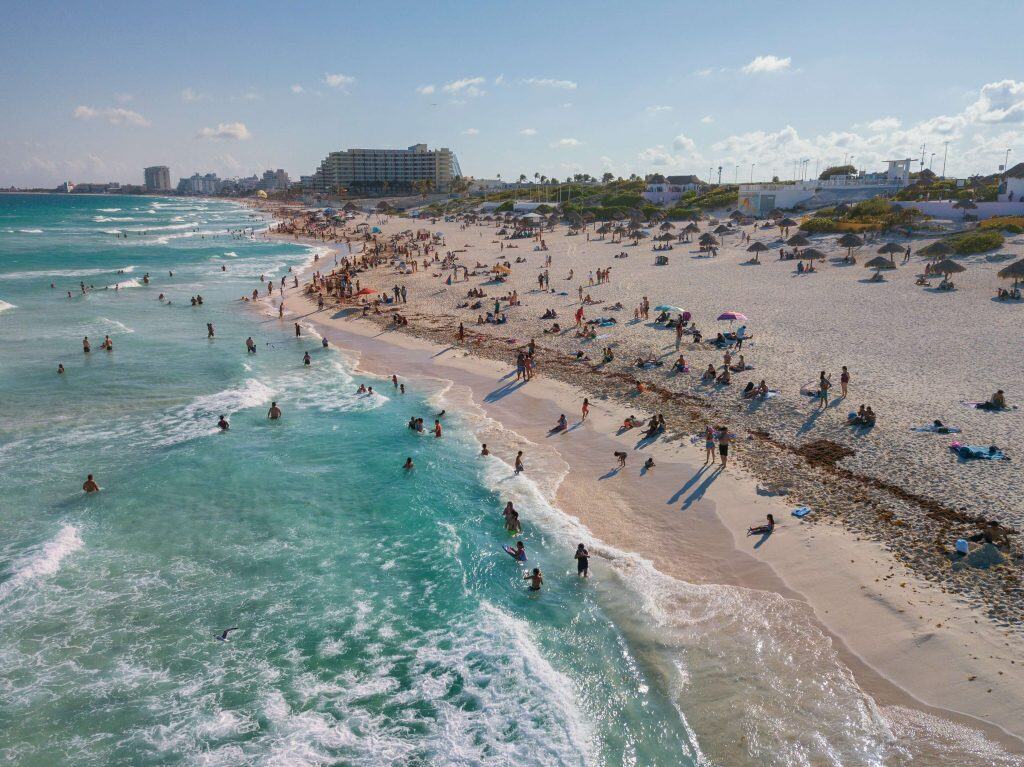 A busy beach scene with people swimming in turquoise water, sunbathing on white sand, and relaxing under umbrellas. Buildings and palm trees are visible in the background under a sunny, blue sky.
