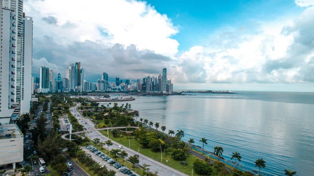 Aerial view of a coastal city with tall skyscrapers, a waterfront highway lined with palm trees, green parks, and calm blue ocean under a partly cloudy sky.